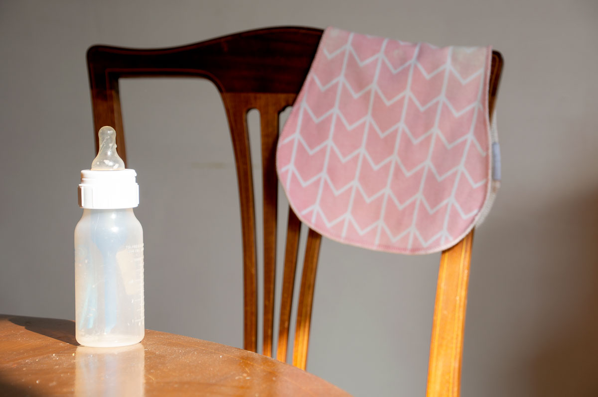 An empty baby bottle sitting on a dining table with a burp cloth draped over a chair in the background.